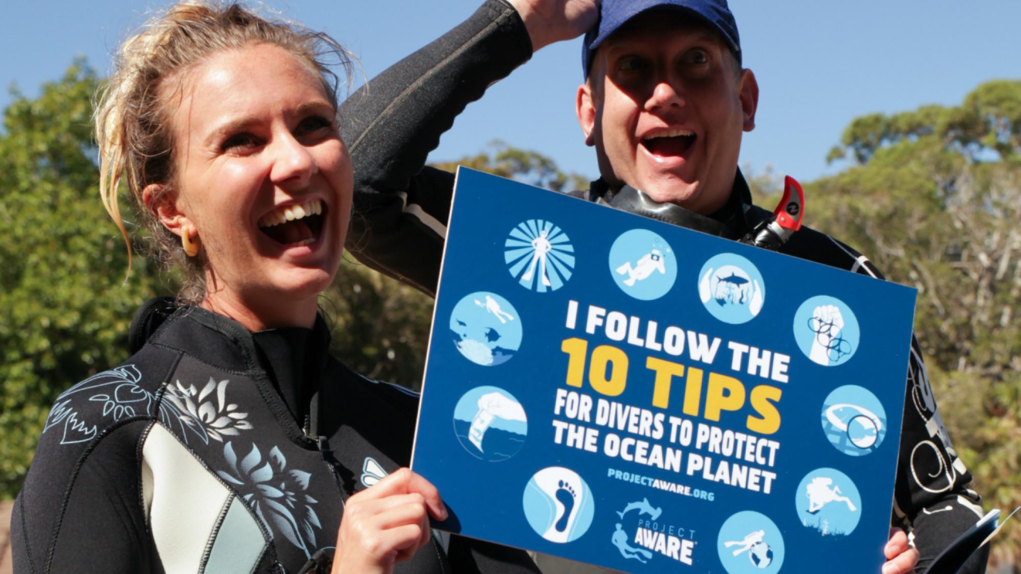 Two scuba divers smiling and holding a sign that says 'I follow the 10 tips for divers to protect the ocean planet' from Project Aware.