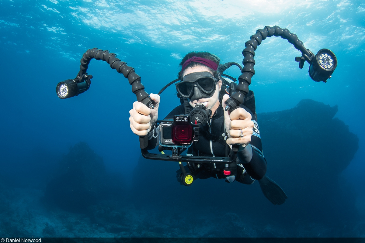 A diver underwater, holding a camera with lights attached, surrounded by blue water.