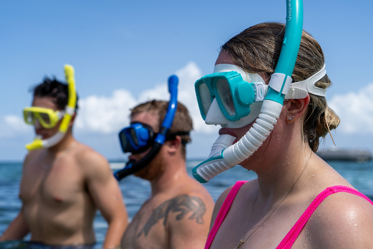 Three snorkelers in shallow water, wearing colorful masks and snorkels, with a clear blue sky and distant clouds in the background.