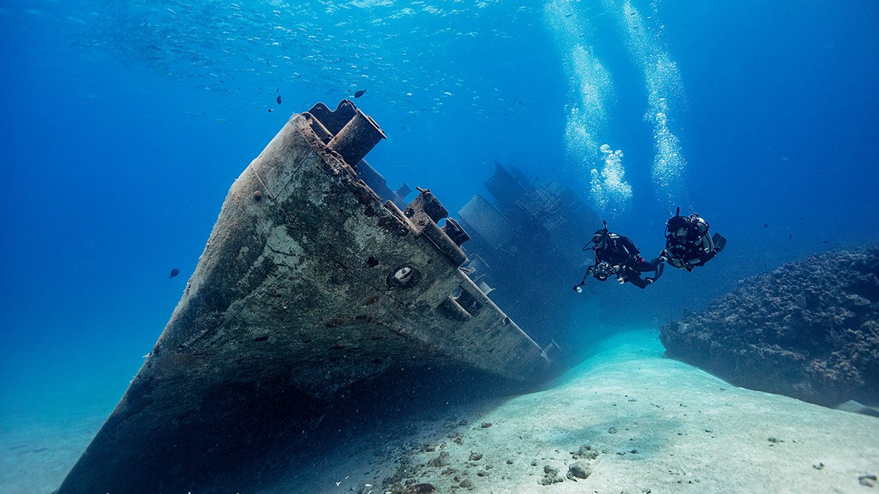 Two scuba divers explore a submerged shipwreck surrounded by clear blue water and small fish.