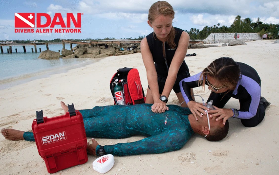 Two divers performing CPR on a submerged individual on a beach, with a red first aid kit nearby.