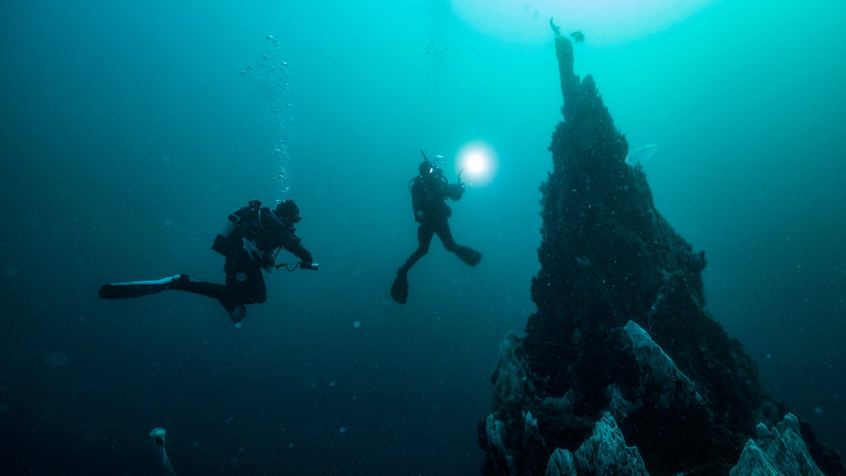 Two scuba divers exploring underwater terrain with a rocky formation in the background and bubbles rising in the water.