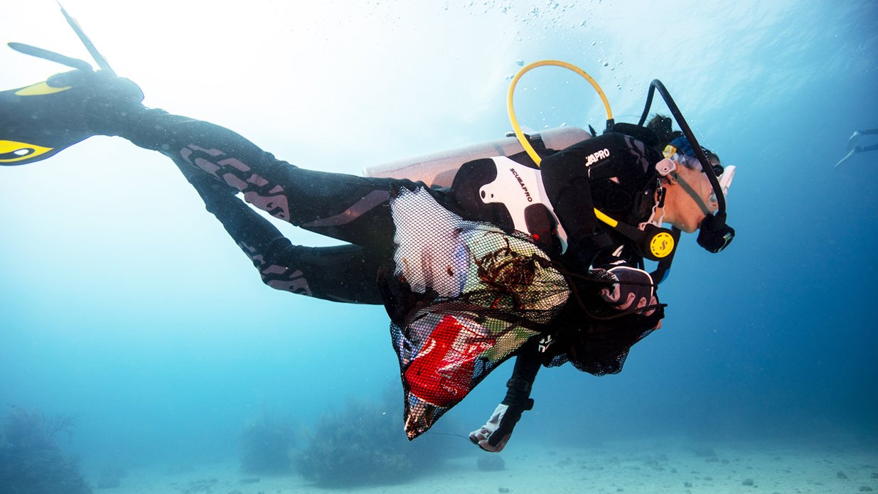 A scuba diver swimming underwater, wearing a dive suit and tank, with a mesh bag filled with items slung over their shoulder.
