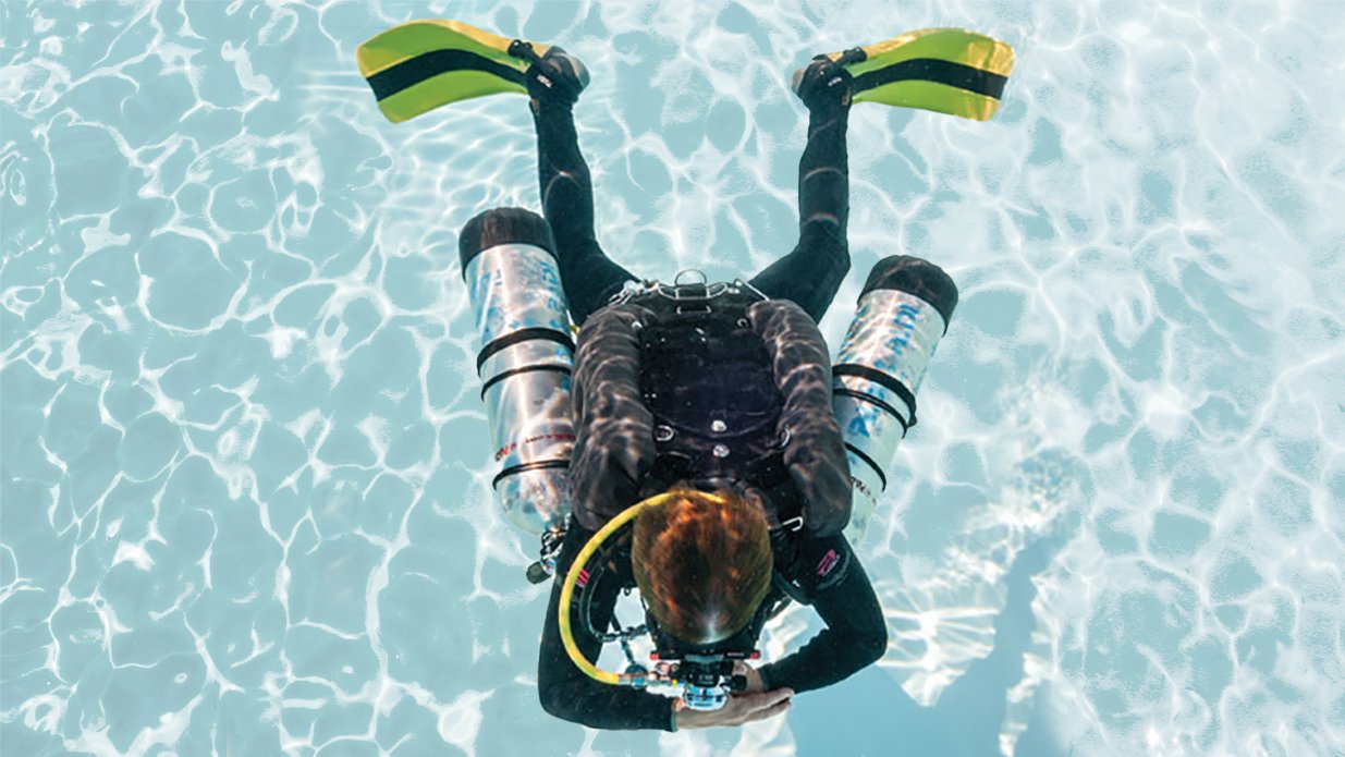 A scuba diver with yellow fins and a black wetsuit is diving underwater, engaged in capturing underwater photography.