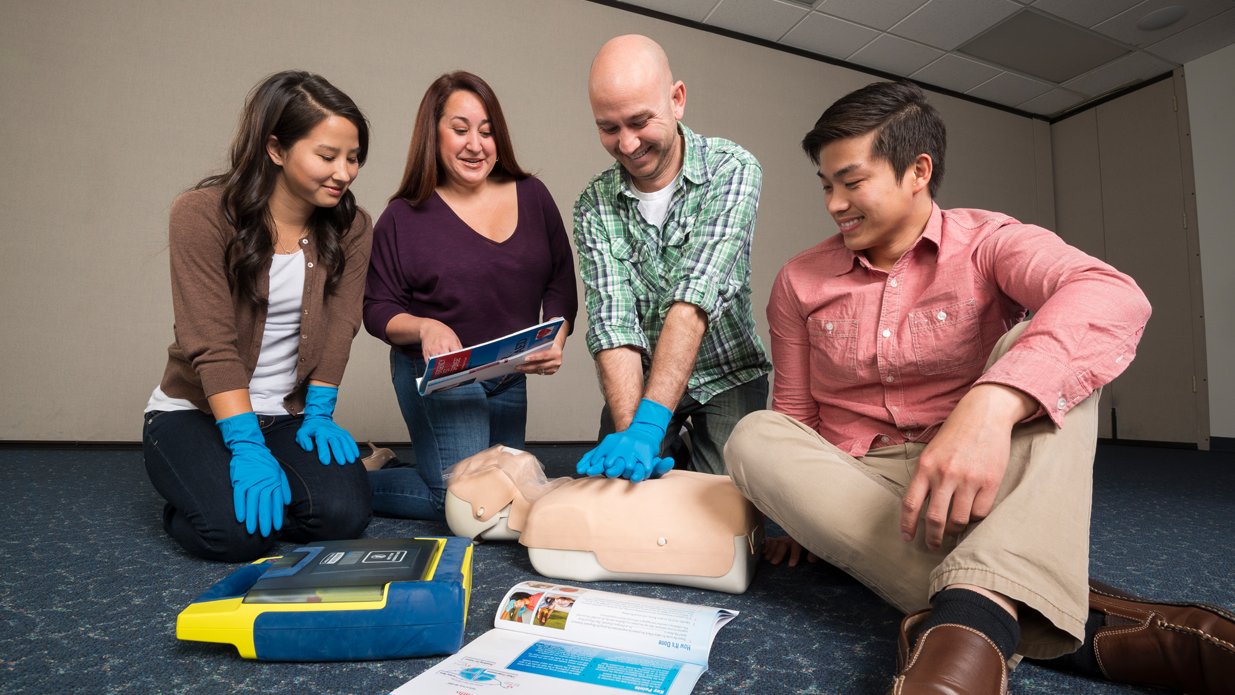 Four individuals learning CPR in a training session, with a CPR mannequin and instructional materials on the floor.