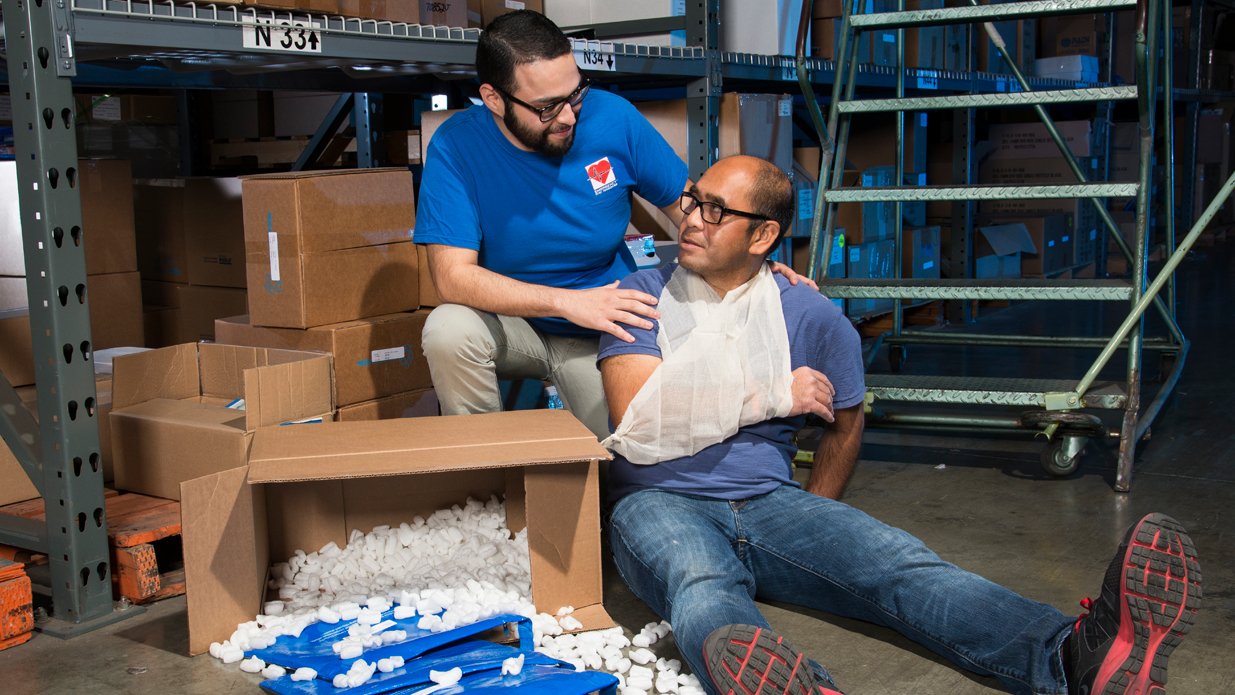 Two men in a warehouse setting; one man is sitting on the floor with a bandaged arm, while another man is providing assistance and support.