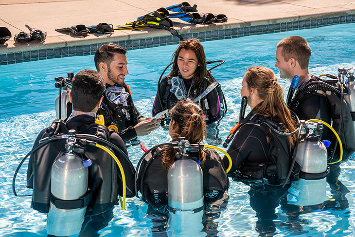 A group of six divers in scuba gear gathering in a swimming pool, with one instructor demonstrating equipment usage.