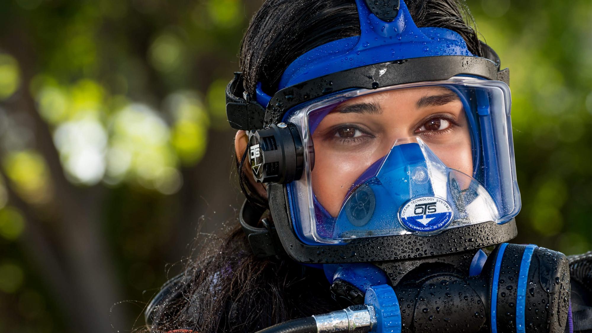 Close-up of a person wearing a blue scuba diving mask and gear, with droplets of water on the mask. The background features lush greenery.