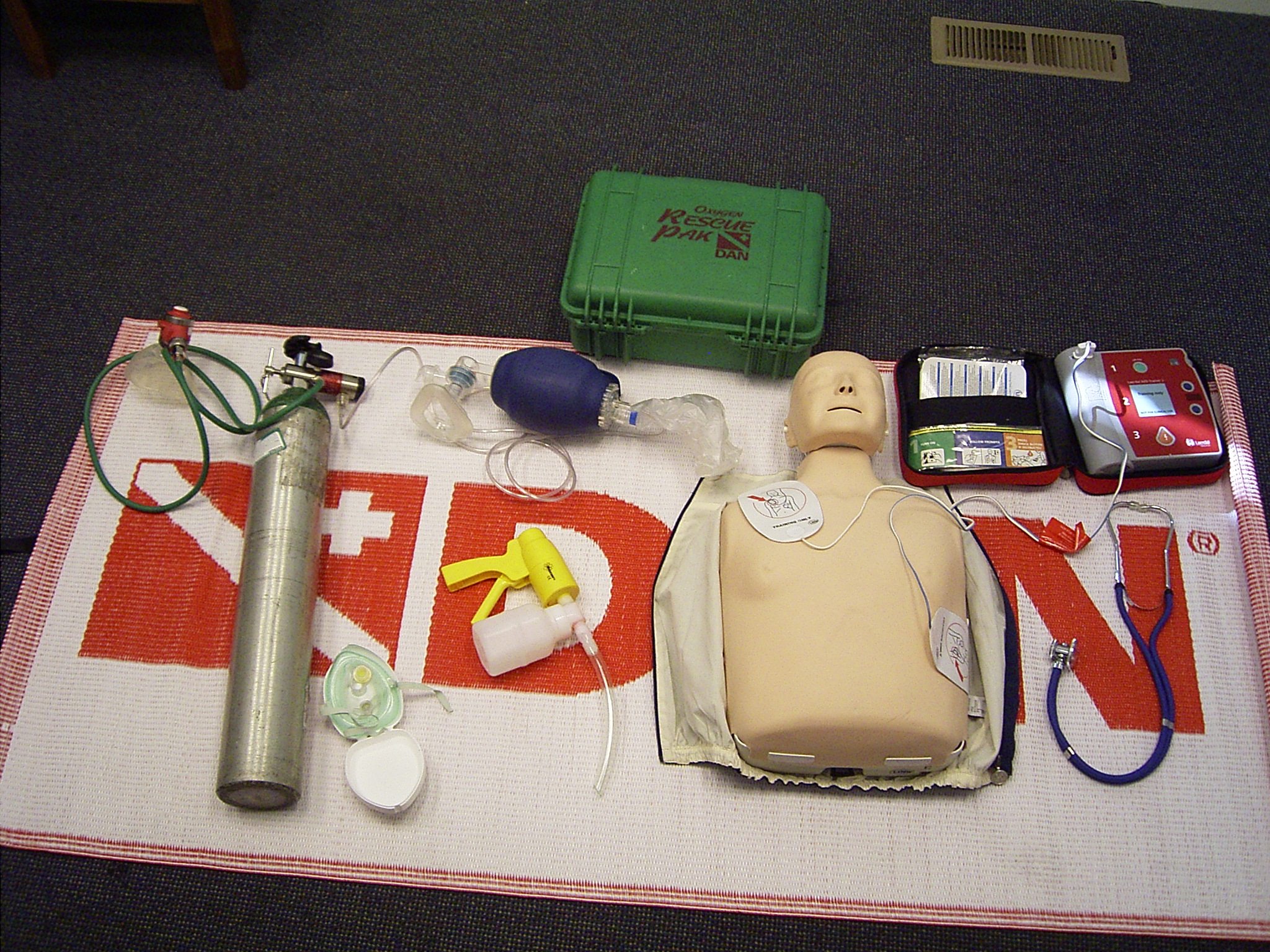 A first aid training setup featuring a CPR mannequin, various medical tools including an oxygen cylinder, a bag-valve mask, and a defibrillator, arranged on a first aid mat.