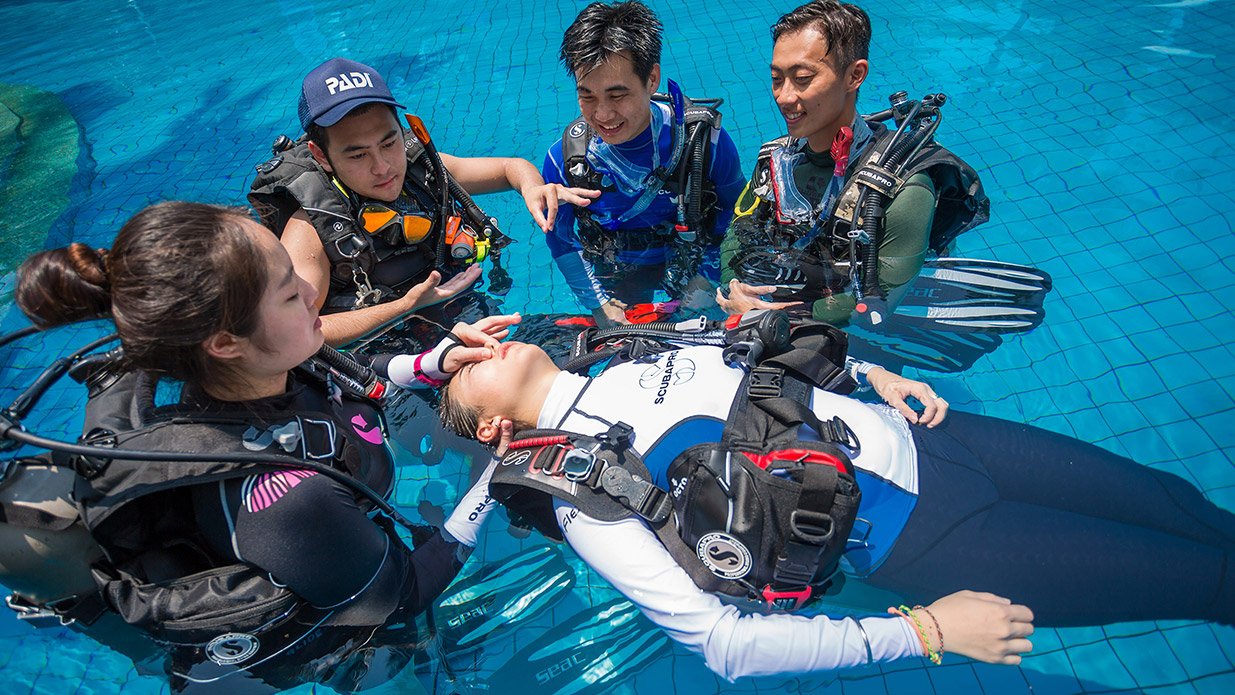 A group of four scuba divers in a swimming pool, with one diver lying back in the water while the others assist and interact with her. They are all wearing diving gear, and the scene captures a training or safety practice.