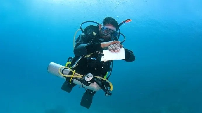 A scuba diver underwater holding a clipboard, surrounded by blue ocean water.
