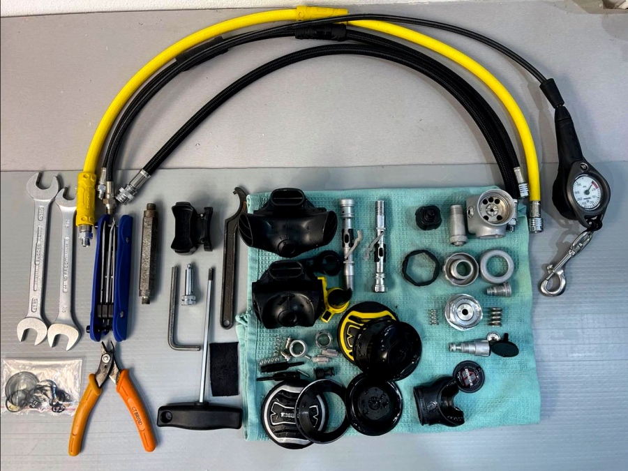 A collection of scuba diving tools and equipment arranged on a surface, including hoses, wrenches, gauges, regulators, and various connectors.