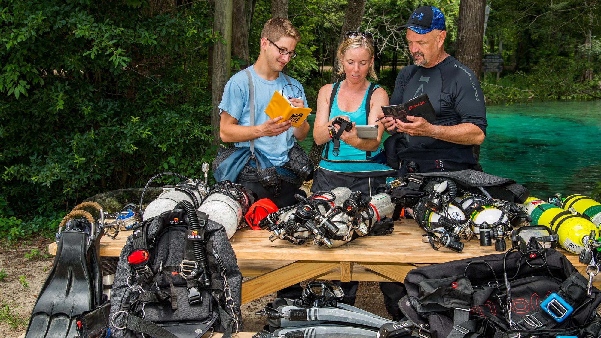 Three divers reviewing equipment manuals and dive guides at a wooden table surrounded by diving gear in a natural setting.