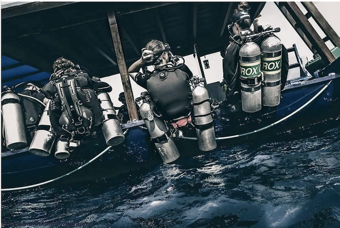 Three divers in scuba gear preparing to enter the water from a boat, with scuba tanks and diving equipment visible.